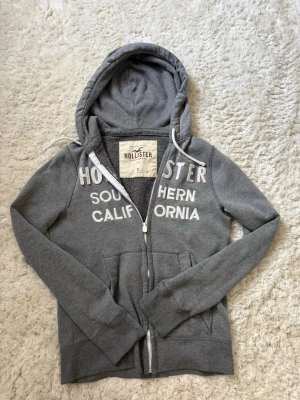 Grå Hollister Zip Hoodie - Säljer min fina hollister hoodie i grå, för att jag inte använder den längre. Tröjan har dragkedja framtill, två fickor och justerbar huva med snören. Den är i storlek S. Den är använd men inte sliten💗 Säljer den för 300kr, skriv bara om ni undrar nåt!!