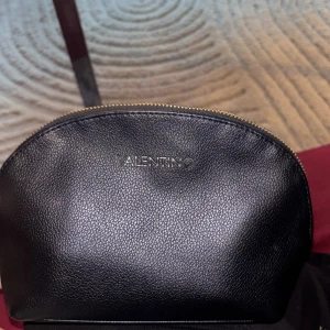 Svart necessär från Valentino - Snygg svart necessär från Valentino i slätt konstläder. Halvmåneformad design med guldfärgad dragkedja upptill och diskret logga framtill. Perfekt för att organisera småsaker eller smink i väskan.