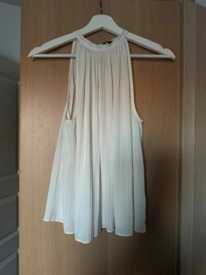 Vit ärmlös linnetopp från Zara - Säljer en snygg vit linnetopp från Zara i storlek S. Toppen är ärmlös med rund halsringning och har ett plisserat, luftigt tyg som ger ett flowy intryck. Perfekt för varma dagar och enkel att styla med jeans eller kjol.
