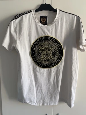 Vit t-shirt från Glorious Gangsta - Vit t-shirt från Glorious Gangsta med stort svart och guldigt broderat emblem på bröstet. Klassisk rund hals, korta ärmar och svarta detaljer längs axlarna. Perfekt för dig som vill sticka ut med en unik och trendig look.
