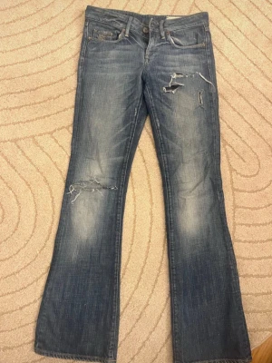 Blå lågmidjade bootcut slitna jeans  - Säljer ett par blå jeans från G-Star RAW med slitningar och hål på låret och knät. Jeansen har bootcut-modell och klassisk femficksdesign. Materialet är denim i bomull och de har en snygg tvättad look.