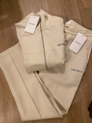 Beige mjukisset från Les Deux - Säljer ett par stilrena beige mjukisbyxor från Les Deux med diskret logga på benet. Byxorna har resår i midjan och är gjorda i ett mjukt och bekvämt material, perfekta för en avslappnad look. Matcha med hoodie för en komplett outfit.