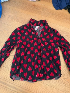 Victoria secret pyjamas-set - Säljer ett svart pyjamas-set med långa ärmar och knäppning framtill. Pyjamasen har ett mönster av röda hjärtan över hela plagget och en bröstficka. Perfekt för mysiga kvällar hemma. Bra skick och inga defekter. Orginal pris runt 900-1000