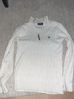 Vit kabelstickad tröja Polo Ralph Lauren - Vit kabelstickad tröja från Polo Ralph Lauren med halv dragkedja och ribbad krage. Tröjan har lång ärm, klassisk logga broderad på bröstet och detaljer i läder på dragkedjan. Perfekt för lager-på-lager och stilren look.