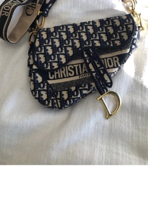 Saddle Bag blå/vit - Ikonisk Saddle Bag i blått och vitt canvasmaterial med det klassiska D-monogrammet. Väskan har guldfärgade metalldetaljer, inklusive ett D-format hänge, och bred axelrem med logotyp. Snygg och trendig design som sticker ut.