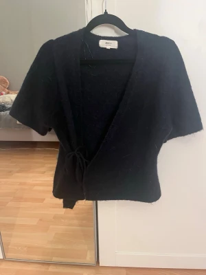 Svart omlottkofta från GinaTricot - Mysig svart omlottkofta från GinaTricot med korta ärmar och knytband i midjan. Koftan har en mjuk, luddig yta och en avslappnad passform som ger en cool och trendig look.