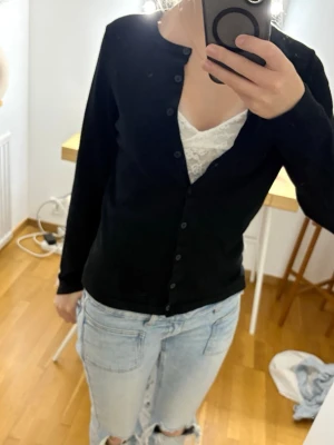 Svart basic kofta med knappar - En enkel svart kofta med långa ärmar och knäppning framtill. Koftan är lätt matcha med jeans eller kjol och är lätt att ha bågot underMaterialet ser mjukt och stretchigt ut, vilket gör den bekväm att bära hela dagen.