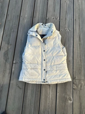 Beige dunväst från Everest - Stilren beige dunväst från Everest med  krage och tryckknappar framtill. Västen har två sidofickor och en innerficka, samt en mjuk vaddering. Perfekt för lager-på-lager och enkel att matcha med olika outfits. Dragkedjans handtag har åkt av (se sista bilden) och en liten fläck framtill (se näst sista bilden) men annars är västen i mycket fint skick!