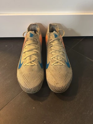 Nike Mercurial orange/blå fotbollsskor - Säljer ett par Nike Mercurial fotbollsskor i orange med blå swoosh och orange detaljer på hälen. Skorna har snörning, låg profil och ovandel i stickat material för bra passform och känsla på planen. Perfekta för dig som vill ha stilrena och snabba skor på matchen.