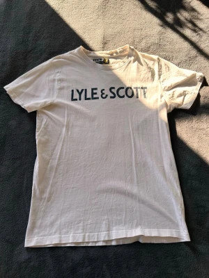 Vit Lyle & Scott t-shirt  - Snygg vit t-shirt från Lyle & Scott med stort svart logotryck över bröstet. Klassisk rund halsringning och rak passform. Tillverkad i mjuk bomull som känns skön mot huden. Perfekt för en avslappnad och stilren look. Nypris 200 Kr. Litet hål nere på ryggen.