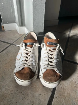 Philippe Model vita och bruna sneakers - Säljer ett par vita sneakers från Philippe Model med bruna mockadetaljer på tån och tungan. Skorna har vita snören, svart detalj på hälen och den klassiska sköldloggan på sidan. Tillverkade i en mix av läder och mocka för en schysst kontrast. Man kan se på bilderna att de är slitna på vissa ställen, därav billiga priset 