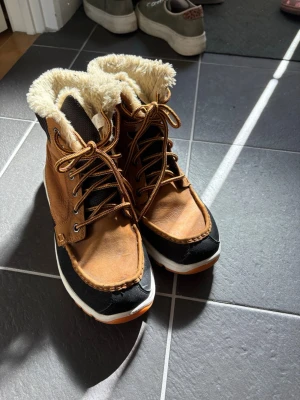 Bruna vinterkängor från Helly Hansen - Säljer ett par bruna vinterkängor från Helly Hansen med svart och vitt detaljer. Kängorna har snörning, grov sula och är fodrade med mjuk, beige fuskpäls för extra värme. Perfekta för kalla dagar och har en robust design. Skorna är helt oanvända. Stått i sko-garderob sedan köp.