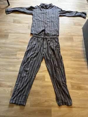 Randig pyjamas i ljusblå och vitt - Säljer en tvådelad pyjamas med skjorta och byxor i ljusblå tyg med vita vertikala ränder. Skjortan har krage och långa ärmar, byxorna har resår i midjan för extra komfort. Perfekt för mysiga kvällar hemma.