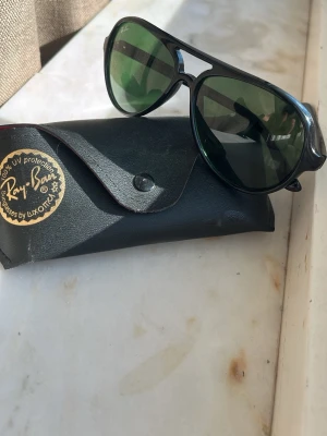 Svarta Ray-Ban pilotbågar solglasögon - Klassiska svarta Ray-Ban solglasögon i pilotmodell med gröntonade glas. Snygg metallbåge och ikonisk design som ger en cool vibe. Kommer med original svart fodral med Ray-Ban-logga. Perfekta för soliga dagar!