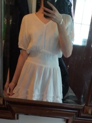 Vit kortklänning från Gina Tricot - Supersöt vit kortklänning från Gina Tricot med v-ringning och smock i midjan. Klänningen är gjord i ett mjukt, strukturerat bomullstyg och har en luftig, flowy passform som känns perfekt för varma dagar.