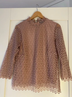 Rosa spetsblus med trekvartsärm - Söt rosa blus i genombruten spets med cirkelmönster och hög hals, Blusen har trekvartsärmar och två klädda knappar nacken för enkel påklädning. Perfekt för dig som gillar romantisk och trendig stil.