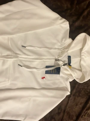 Vit hoodie från Polo Ralph Lauren - Snygg vit hoodie från Polo Ralph Lauren med dragkedja och huva. Klassisk röd broderad logga på bröstet och justerbara snören i huvan. Perfekt för en clean och stilren look. Materialet känns mjukt och bekvämt.