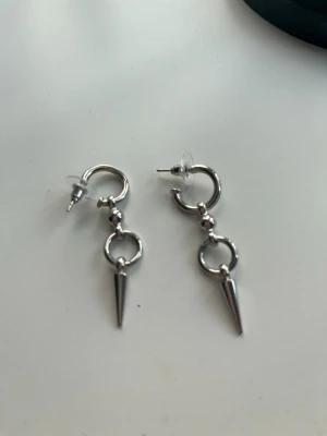 Silvriga örhängen med konformad hängare - Säljer ett par silvriga örhängen med rund ring och en konformad hängande detalj. Örhängena har push-back lås i plast och en stilren, edgy design som passar dig som gillar att sticka ut. Perfekta för att ge din look en cool touch.