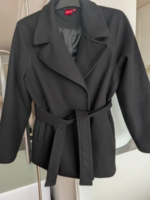 Svart jacka med skärp från RYVLS - Stilren svart jacka från RYVLS (Kids Brand Store) med klassisk krage och knytband i midjan samt två tryckknappar. Kappan har två fickor framtill och är helfodrad för extra komfort. Perfekt för dig som vill ha en enkel men snygg look till vår eller höst. Stlk 158/164 men passar XS