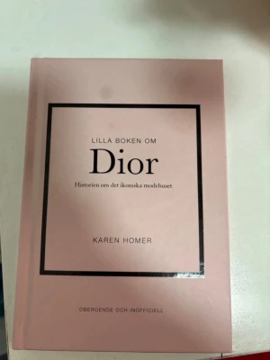 Lilla boken om Dior av Karen Homer - En stilren, ljusrosa bok med titeln 'Lilla boken om Dior' av Karen Homer. Boken handlar om historien bakom det ikoniska modehuset Dior och har ett elegant omslag med svart ram och text. Perfekt för modeintresserade. Har två små fläckar som man kan se på bilden men inget som märks av 