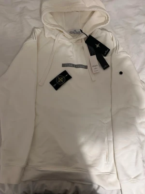 Vit hoodie från Stone Island - Snygg vit hoodie från Stone Island med klassisk huva och dragsnören. Hoodien har en stilren design med logotyp på bröstet och den ikoniska avtagbara patchen på ärmen. Tillverkad i mjukt bomullsmaterial för maximal komfort.