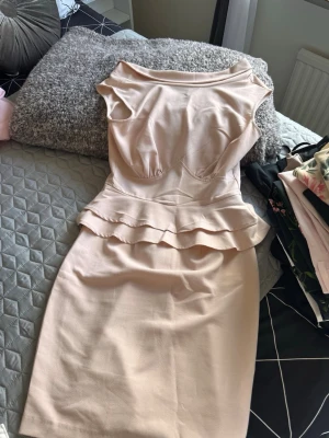 Ljusbeige peplumklänning med volang - Elegant ljusbeige klänning med peplumvolang i midjan och figurnära passform. Klänningen har rund halsringning, är ärmlös och har dragkedja i ryggen. Materialet känns som stretchig polyesterblandning och längden går till knäna.