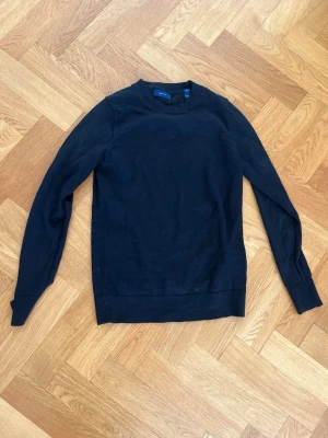 Svart långärmad tröja från Gant - En stilren svart tröja från Gant med rund hals och långa ärmar. Tröjan har en enkel, klassisk design och är tillverkad i mjukt material som passar perfekt till alla outfits. Ribbstickade muddar vid ärmslut och nederkant ger en snygg finish.