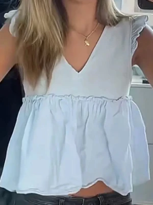 Ljusblå volangblus - Supersöt Sthlmstil ljusblå blus med v-ringad hals och volangdetaljer. Ärmlös modell med små volanger på axlarna och luftig passform. Perfekt till jeans eller kjol för en fräsch look.