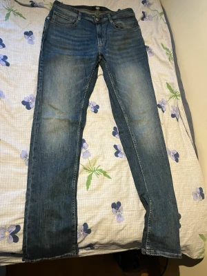 Blå raka jeans från KappAhl - Säljer ett par klassiska blå jeans från KappAhl med rak passform och fem fickor. Jeansen har en snygg tvätt med slitningar på låren och knäna. Perfekta för en avslappnad och trendig look.