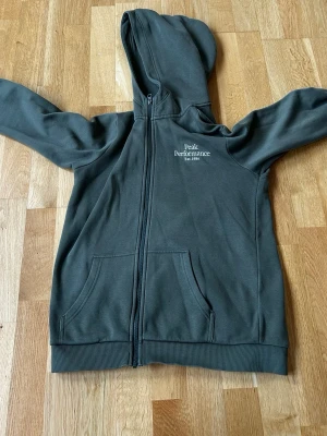 Mörkgrön hoodie Peak Performance - Säljer en mörkgrön hoodie från Peak Performance med dragkedja framtill och två stora fickor. Märket är broderat i vitt på bröstet. Hoodien har ribbade muddar och en klassisk huva. Perfekt för en avslappnad och sportig stil.