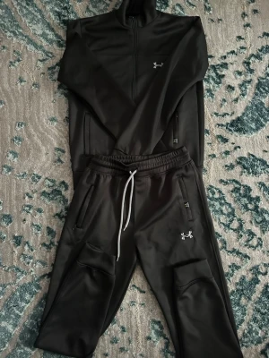 Svarta mjukis sett från Under Armour priset går att diskutera - Säljer ett par svart gråa mjukis sett från Under Armour med vita dragsnören och resår i midjan. Byxorna har två sidofickor med dragkedja och en liten logga på vänster ben och det är samma med tröjan. Sköna och stilrena, perfekta för chill eller träning. Priset kan diskuteras!!