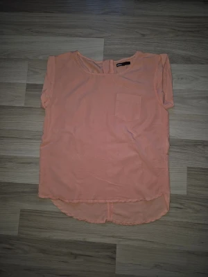 Korallfärgad blus med ficka - Säljer en Korallfärgad blus med rund halsringning och korta ärmar. Toppen har en liten bröstficka och rundad nederkant som är något längre bak. Perfekt för en avslappnad och trendig look.