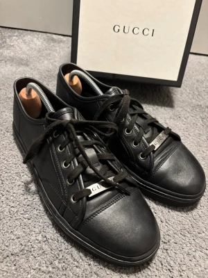 Gucci Ace all black leather - Snygga svarta sneakers från Gucci i slätt läder med svarta skosnören och metallbricka med Gucci-logga. Klassisk låg modell med rund tå och diskreta sömmar. Perfekta för dig som gillar stilrena och exklusiva skor.