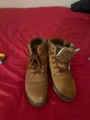 Bruna kängor med snörning och foder - Snygga bruna kängor med grov sula och snörning. Insidan är fodrad med mjukt material för extra värme. Kängorna har metallhål för snörena och kontrastsömmar längs sulan. Perfekta för kalla dagar och ger en cool look till din outfit. För ålder 13 och upp