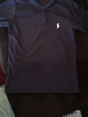 Marinblå pikétröja från Polo Ralph Lauren - Säljer en klassisk marinblå pikétröja från Polo Ralph Lauren med vit broderad logga på bröstet. Tröjan har krage, knappar vid halsen och är tillverkad i mjuk bomull. Perfekt för en clean och stilren look.