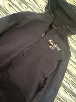 Svart hoodie från Napapijri - Säljer en svart hoodie från Napapijri med dragkedja och tryckt logga på bröstet. Hoodien har huva och är tillverkad i mjukt material som känns skönt mot huden. Perfekt för dig som gillar streetwear och vill ha en stilren look.