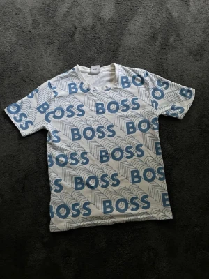 Vit och blå BOSS t-shirt - Säljer en vit t-shirt från Hugo Boss med blått BOSS-tryck över hela tröjan. T-shirten har rund halsringning och korta ärmar. Materialet känns mjukt och skönt, perfekt för en avslappnad stil.