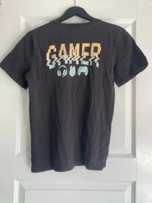 Svart t-shirt med gamer-tryck - Svart t-shirt med coolt gamer-tryck på bröstet i pixelstil. Trycket visar texten 'GAMER' med vågig effekt och ikoner för headset, mus och handkontroll. T-shirten har rund hals och korta ärmar, perfekt för dig som gillar gaming och vill visa det med stil.