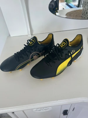 Svarta och gula Puma fotbollsskor - Snygga fotbollsskor från Puma i svart och gult med gulddetaljer och klassisk logga på sidan. Skorna har snörning, låg profil och dobbar för spel på gräs. Insidan är gul och designen är inspirerad av King-modellen med signaturdetaljer. Använda 1-2 ggr