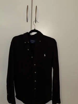 Svart skjorta från Polo Ralph Lauren - Klassisk svart skjorta från Polo Ralph Lauren med knappstängning och ikonisk broderad logga på bröstet. Skjortan har lång ärm och är tillverkad i ett lätt bomullsmaterial som känns skönt mot huden. Perfekt för en clean och stilren look.