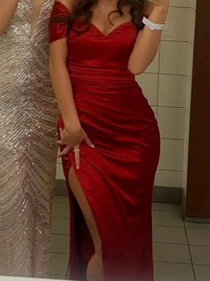 Röd balklänning med slits - Elegant röd långklänning i glansigt material med offshoulder och figurnära passform. Klänningen har en hög slits på ena sidan och vackra draperingar längs midjan och höften. Perfekt för bal eller festliga tillfällen. Nypris: 3700kr