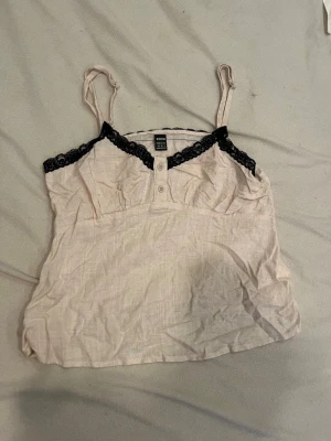 Ljusbeige linnetopp med spetsdetaljer - Söt ljusbeige linnetopp med svarta spetsdetaljer längs urringningen och tunna axelband. Toppen har knappar framtill och är tillverkad i ett lätt och luftigt material, perfekt för varma sommar dagar.