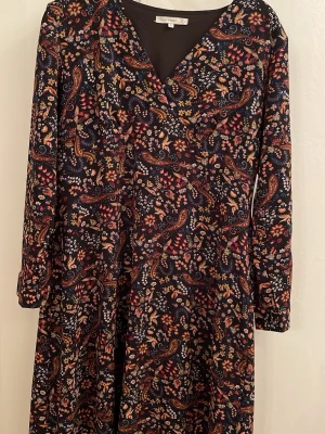 Mönstrad klänning från Culture - Säljer en långärmad klänning från Culture med mörk botten och färgglatt paisleymönster i rött, gult, blått och vitt. Klänningen har v-ringning och mjukt, följsamt tyg som ger en avslappnad passform. Perfekt för dig som gillar bohemiska vibes och unika prints.