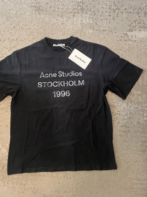 Svart Acne Studios t-shirt - Snygg svart t-shirt från Acne Studios med tryck 'Acne Studios STOCKHOLM 1996' på bröstet. Klassisk passform med rund hals och korta ärmar. Tillverkad i mjuk bomull för en skön känsla. Perfekt till jeans eller shorts.