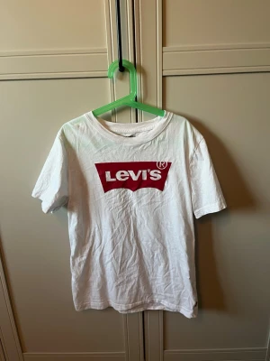 Vit Levi's t-shirt med röd logga - Säljer en klassisk vit t-shirt från Levi's med den ikoniska röda loggan tryckt på bröstet. T-shirten har rund hals och korta ärmar. Tillverkad i mjuk bomull och passar perfekt till jeans eller shorts.