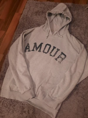 Grå hoodie med tryck 'AMOUR' - Säljer en ljusgrå hoodie med huva och dragsko. På framsidan står det 'AMOUR' i svart, slitet tryck och på ryggen finns en stjärna i samma stil. Hoodien har ribbade muddar vid ärmar och nederkant. Perfekt för en chill och avslappnad look.