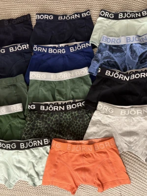 16 Par Björn Borg Boxerkalsonger för Barn - Ett stort paket med 16 par Björn Borg boxerkalsonger för barn! ✨Samtliga är i storlek 110-116 och materialet på alla är 95% bomull & 5% elastan. Olika färger och mönster:                         grön, mörkgrön, olivgrön/khaki, marinblå/mörkblå, blå, ljusblå, 2 x mintgrön, 2 x svart, 2 x grå, orange samt två par med grönt/svart och blått mönster. Midjan har resår och Björn Borg-tryck. Alla är i mycket fint använt skick, utan några defekter. Ett par svarta och ett par gråa syns ej på bild! 
