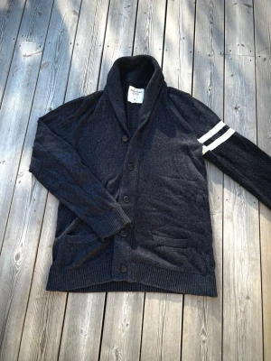 Abercrombie & Fitch cardigan - En riktigt fräsch cardigan perfekt till våren och sommarkvällen i märket Abercrombie & Fitch i en snygg mörkblå färg. Den är stickad i ett skönt material och har vita detaljer som ränderna på ärmen. Den är i bra skick men inga defekter. Storleken är Xl och mått finns i bilderna men kom gärna Pm för fler frågor.