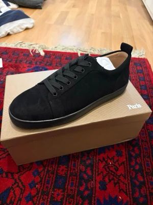 Svarta sneakers från Christian Louboutin - Stilrena svarta sneakers i mocka från Christian Louboutin med klassisk röd sula och svarta skosnören. Låg modell med diskreta detaljer och bekväm passform. Perfekta för dig som vill ha en clean look med exklusiv känsla.