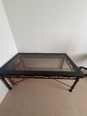  Bord  - Säljer ett stilrent soffbord med svart metallram och rektangulär glasskiva. Bordet har dekorativa detaljer på benen och en stabil konstruktion. Perfekt som vardagsrumsbord för dig som gillar modern och industriell stil.